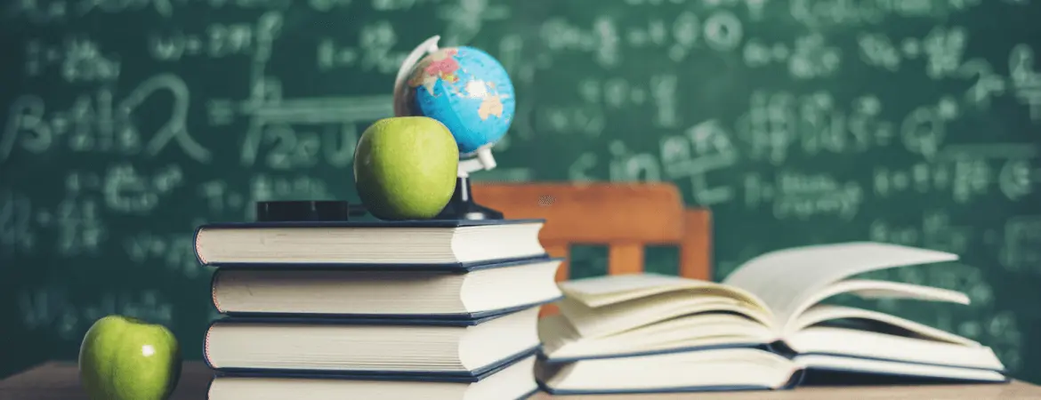 Stack of books with apples and a globe in front of a chalkboard.